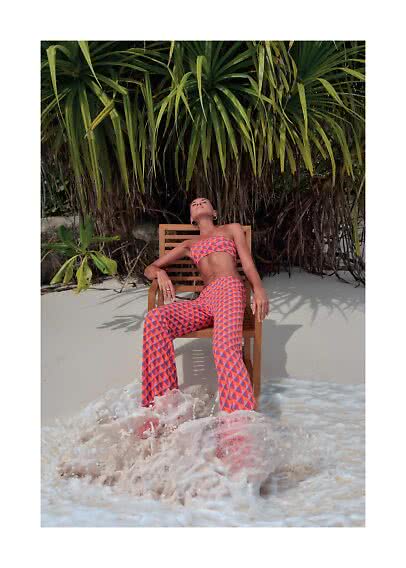 Woman in patterned bandeau top and pants sits on a chair on the beach with waves at her feet