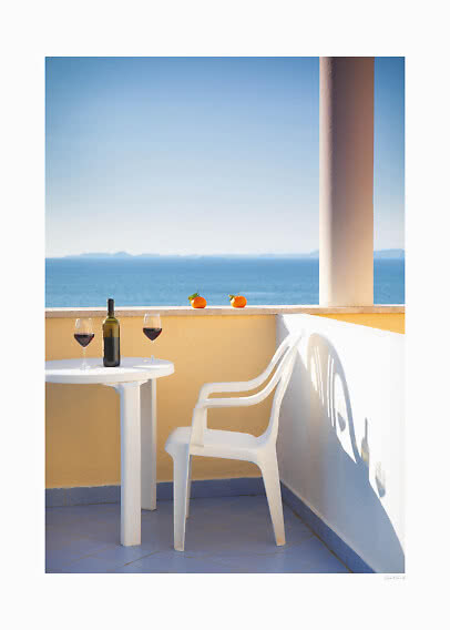 Sunny balcony with white chair and table, wine bottle, two glasses of red wine and sea view