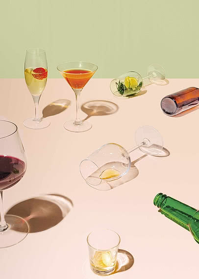 Tisch mit Cocktailgläsern, Wein, Shots und Flaschen auf pastellfarbener Fläche im Sonnenlicht