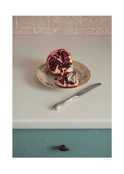 Photograph of an open pomegranate on a floral vintage saucer with a silver knife, on a pale kitchen counter