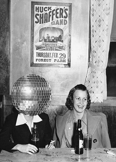 Et svart-hvitt-fotografi av to kvinner ved et barbord med ølflasker og et glass, der hun til venstre har en discokule i stedet for hode