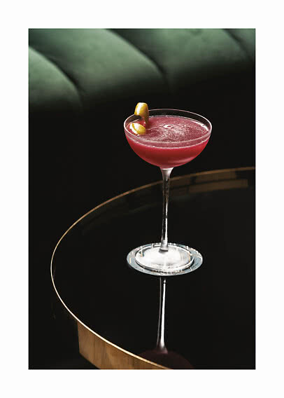 Cocktail rosso in coppa con scorza di limone su tavolo nero davanti a divano verde scuro