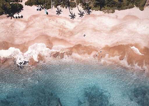 Luftfoto af sandstrand med bølger, palmer og parasoller ved kysten
