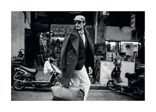 Black-and-white photo of stylish man in coat, cap and sunglasses carrying a large leather bag