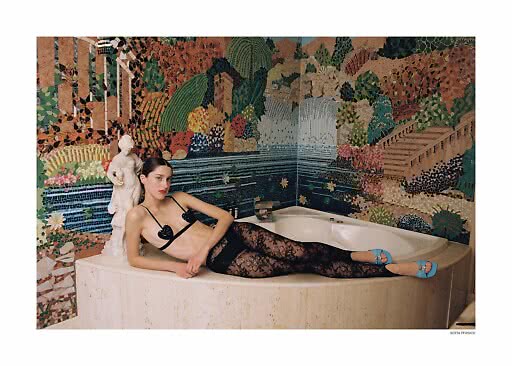 Model in schwarzer Spitzenwäsche und blauen Absatzschuhen an einer Badewanne vor bunter Mosaikwand