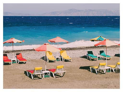 Bunte Sonnenschirme und Liegen am Strand vor türkisblauem Meer mit Bergen im Hintergrund