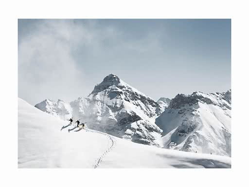 Drei Skifahrer gehen über einen verschneiten Hang vor hohen, schneebedeckten Gipfeln.