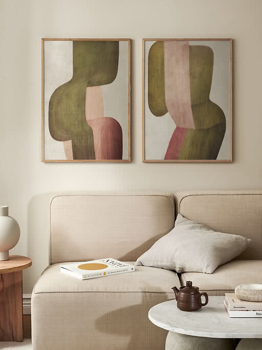 Deux affiches abstraites encadrées au-dessus d’un canapé beige, formes vertes, roses et brunes