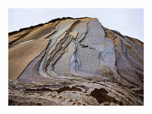 Eine gefaltete Felswand in Sand, Lilagrau und Rost, die Bänder diagonal, oben ein schmales Grasband