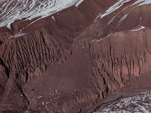 Rust-red Himalayan mountain slope with fine eroded channels and thin streaks of snow along the ridgelines